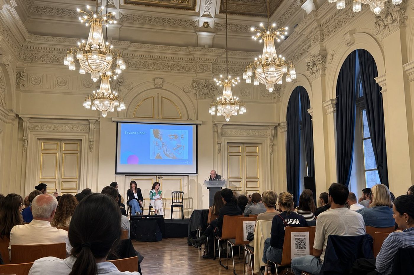 Conference in a gilded hall with chandeliers, audience facing the speakers